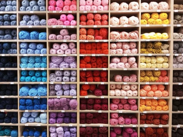 Oke Maisie Moo yarn wall — wooden cubby shelves stacked with hand-picked balls of wool arranged by colour, from blues and purples through reds, pinks and creams to yellows and oranges